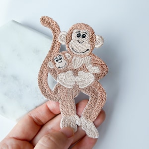 May include: Embroidered patch of two brown monkeys hugging. The larger monkey is holding the smaller monkey in its arms. Both monkeys have black eyes and a light brown face.