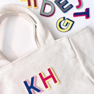 Puede incluir: Un bolso de mano de lona color crema con las letras KH en parches de fieltro rosa, azul y amarillo. Alrededor del bolso hay otras letras de fieltro individuales dispersas en varios colores, incluyendo amarillo, verde, gris, rosa y azul.