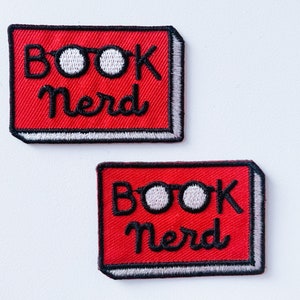 May include: Two red iron-on patches with white stitching. Each patch features the text "Book Nerd" with a pair of glasses in between the words.