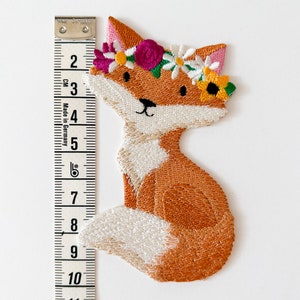 May include: Embroidered patch of a seated fox with orange and white fur, wearing a floral crown with pink, white, and yellow flowers. The patch is approximately 4 inches tall, measured with a tape measure.
