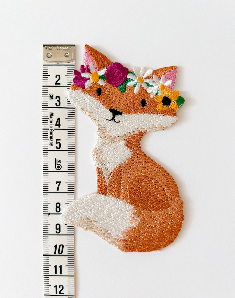 May include: Embroidered patch of a seated fox with orange and white fur, wearing a floral crown with pink, white, and yellow flowers. The patch is approximately 4 inches tall, measured with a tape measure.