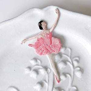 Puede incluir: Un parche bordado de bailarina rosa y blanco con pelo negro sobre un fondo blanco.