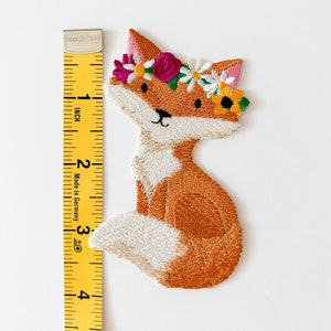May include: Embroidered patch of a fox wearing a floral crown. The fox is orange and white, with a black nose and eyes. The floral crown includes pink, yellow, and white flowers. The patch measures approximately 3 inches in height.