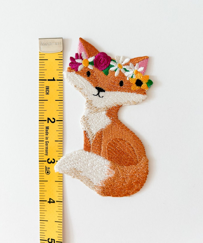May include: Embroidered patch of a fox wearing a floral crown. The fox is orange and white, with a black nose and eyes. The floral crown includes pink, yellow, and white flowers. The patch measures approximately 3 inches in height.