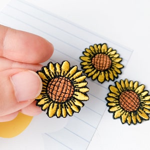 Könnte beinhalten: Drei gelbe gestickte Sonnenblumen-Patches mit braunen Zentren. Die Patches befinden sich auf einem weißen, linierten Hintergrund.