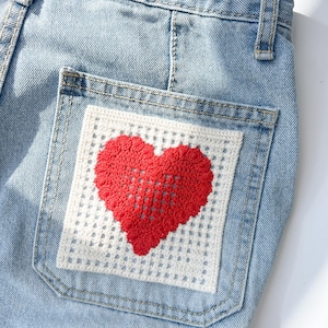 Puede incluir: Vaqueros de mezclilla azul claro con un parche de ganchillo blanco con un diseño de corazón rojo en el bolsillo trasero. El corazón está colocado en el centro del parche, agregando un elemento decorativo a los jeans.