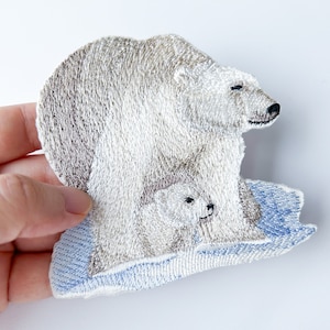 Puede incluir: Parche bordado de un oso polar y una cría de pie sobre un témpano de hielo azul. El oso polar es blanco con la nariz marrón y la cría es blanca.