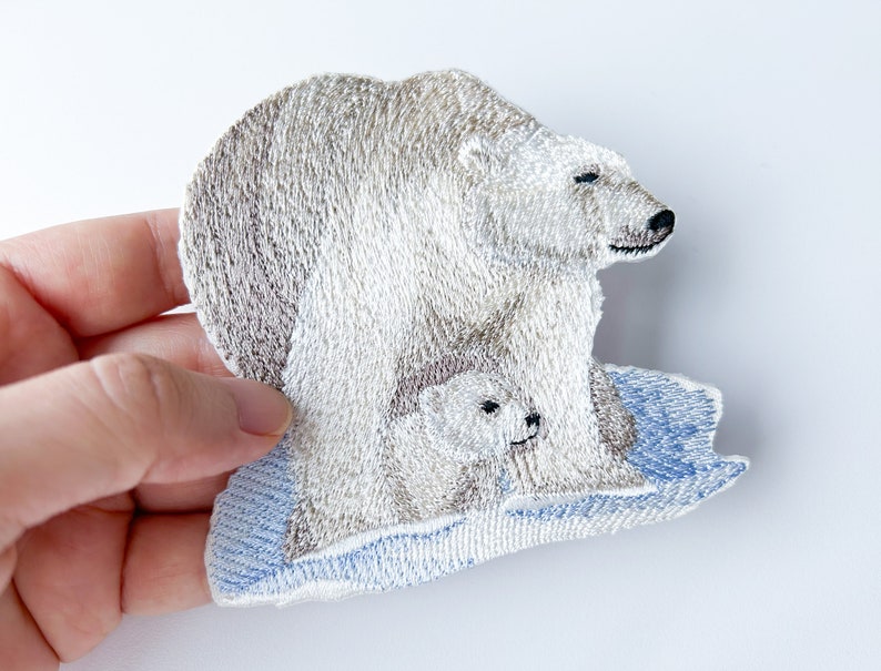 May include: Embroidered patch of a polar bear and cub standing on a blue ice floe. The polar bear is white with a brown nose and the cub is white.