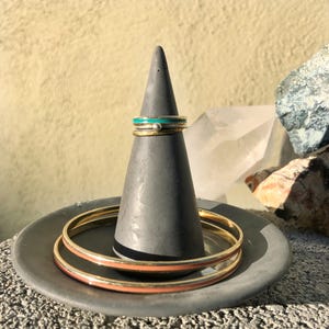 May include: A set of three gold rings with colourful enamel accents. The rings are displayed on a grey concrete ring holder. The rings are stacked on top of each other, with the turquoise ring on top.