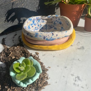 May include: A stack of three ceramic dishes in pastel colors. The top dish is speckled with blue and purple on a cream background. The middle dish is pink, and the bottom dish is yellow. A small succulent sits in a light blue hand-shaped planter.