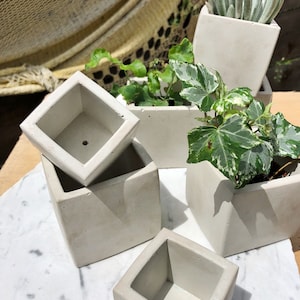 May include: Set of four concrete planters in various sizes and shapes. The planters are a light grey colour and have a rough, textured surface. The planters are empty and ready to be filled with plants.