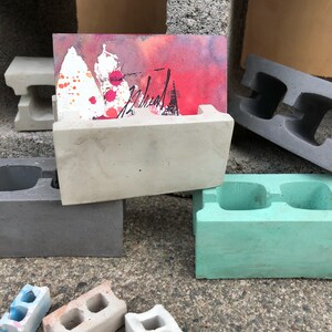 May include: A set of small concrete blocks in various colors, including gray, green, and white. Some blocks have two holes, while others have one. A card holder made of concrete is holding a card with a watercolor painting of a red and white abstract design. The card is signed "Michael".