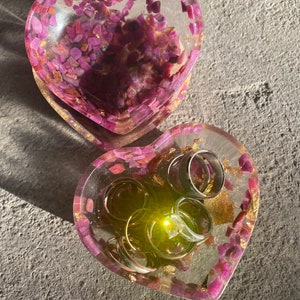 May include: Two heart-shaped resin trinket dishes with pink and gold flecks. The larger dish is empty, while the smaller dish contains several silver rings.