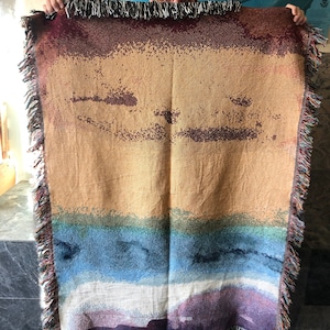 May include: A woven tapestry throw blanket with a colorful abstract design featuring shades of brown, blue, white, and purple. The blanket has a fringed edge.