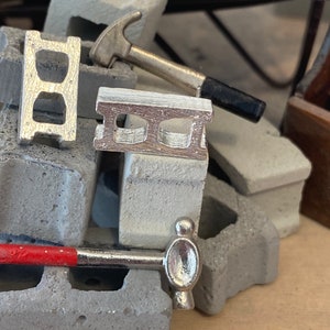May include: A miniature construction set with two silver toy hammers and three concrete blocks. The blocks are made of a light gray material and have a textured surface. The hammers are made of silver metal and have a black handle.