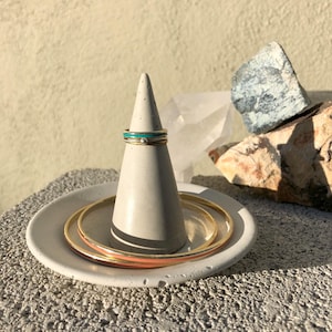 Peut inclure: Un ensemble de bagues en or avec une bande en émail turquoise et blanc sur un porte-bague en béton. Les bagues sont présentées sur un plat en béton blanc avec deux bracelets en or.