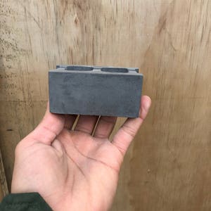 May include: A gray concrete block with two holes in the center. The block is about 3 inches long and 2 inches wide.
