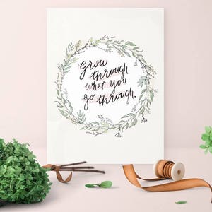 May include: A white print with the text "Grow through what you go through" in a floral wreath. A bundle of green foliage, a spool of ribbon, and a small vase with greenery are on a light pink surface.