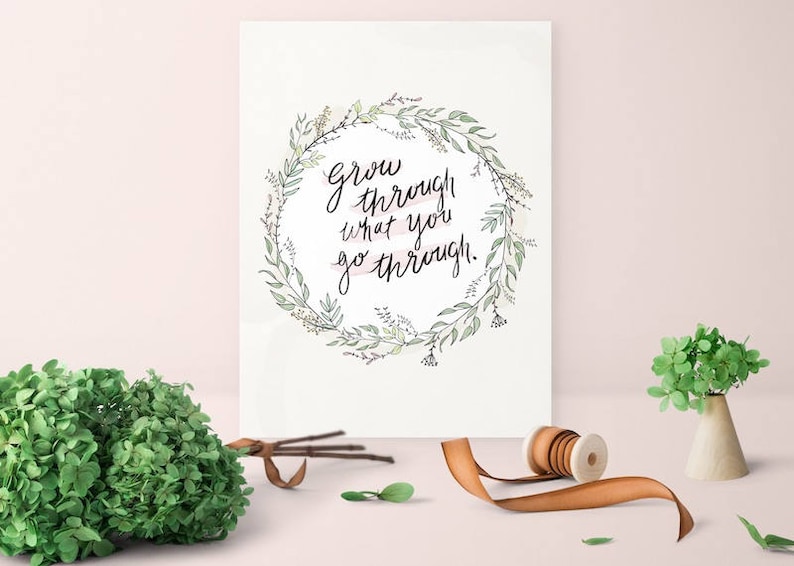 May include: A white print with the text "Grow through what you go through" in a floral wreath. A bundle of green foliage, a spool of ribbon, and a small vase with greenery are on a light pink surface.