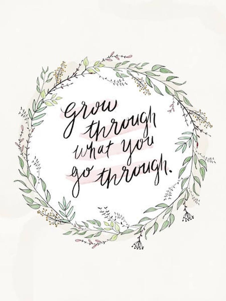May include: A watercolour print with the handwritten phrase "grow through what you go through" in black script. The text is encircled by a delicate wreath of green and lavender leaves and sprigs, set against a soft, off-white background.