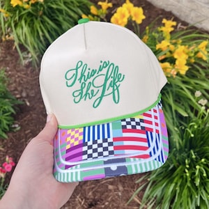 Puede incluir: Gorra de camionero color crema con la frase bordada "This is the Life" en verde. La gorra presenta un colorido patrón geométrico en la visera con un ribete verde.