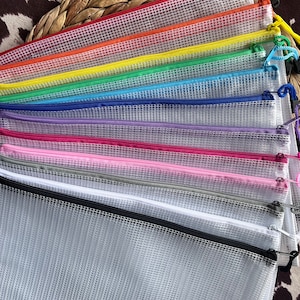 K&ouml;nnte beinhalten: Eine Sammlung farbenfroher Mesh-Rei&szlig;verschlusstaschen in verschiedenen Farben, darunter Rot, Orange, Gelb, Gr&uuml;n, Blau, Lila, Rosa, Grau, Wei&szlig; und Schwarz. Jede Tasche hat einen passenden Rei&szlig;verschluss und eine kleine Schlaufe. Sie liegen auf einer strukturierten Oberfl&auml;che.