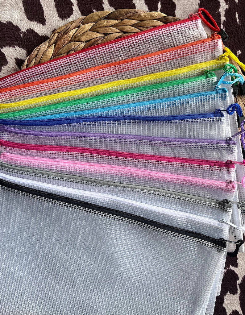K&ouml;nnte beinhalten: Eine Sammlung farbenfroher Mesh-Rei&szlig;verschlusstaschen in verschiedenen Farben, darunter Rot, Orange, Gelb, Gr&uuml;n, Blau, Lila, Rosa, Grau, Wei&szlig; und Schwarz. Jede Tasche hat einen passenden Rei&szlig;verschluss und eine kleine Schlaufe. Sie liegen auf einer strukturierten Oberfl&auml;che.