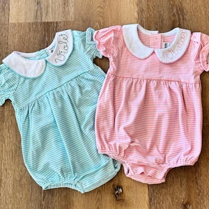 Puede incluir: Dos peleles de bebé, uno celeste y otro rosa, con cuellos blancos y mangas cortas. Cada pelele tiene rayas horizontales y el nombre de un niño bordado en el cuello. Los peleles están sobre una superficie de madera.