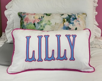 May include: White rectangular cushion with the name "LILLY" in light blue letters with a pink outline. The cushion has a pink border and is on a white bed with other cushions in the background.