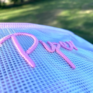 K&ouml;nnte beinhalten: Nahaufnahme einer wei&szlig;en Mesh-M&uuml;tze mit einem Farbverlauf von Hellblau zu Rosa. Das Wort "Purr" ist in einem leuchtenden rosa Schriftzug auf die Vorderseite der M&uuml;tze gestickt. Der Hintergrund ist verschwommen und zeigt gr&uuml;nes Gras und B&auml;ume.