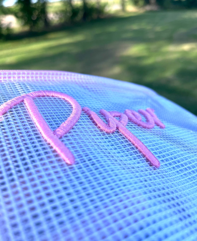 K&ouml;nnte beinhalten: Nahaufnahme einer wei&szlig;en Mesh-M&uuml;tze mit einem Farbverlauf von Hellblau zu Rosa. Das Wort "Purr" ist in einem leuchtenden rosa Schriftzug auf die Vorderseite der M&uuml;tze gestickt. Der Hintergrund ist verschwommen und zeigt gr&uuml;nes Gras und B&auml;ume.