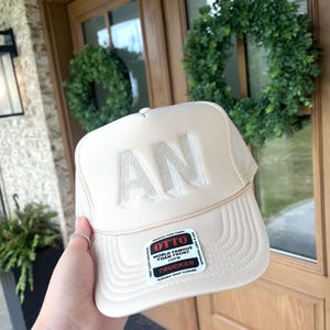 Peut inclure: Casquette de camionneur beige avec les lettres "AN" brodées sur le devant. Le chapeau a un dos en filet et une visière incurvée. Une étiquette sur le chapeau indique "OTTO WORLD FAMOUS FOAM FRONT TRUCKER".