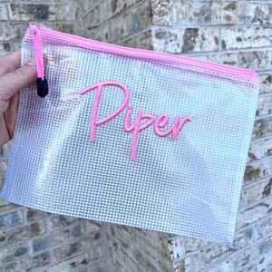 K&ouml;nnte beinhalten: Eine transparente, mit Rei&szlig;verschluss versehene Netztasche mit einem rosa Rei&szlig;verschluss und dem Namen "Piper" in Rosa gestickt. Die Tasche wird vor einer Backsteinmauer gehalten.