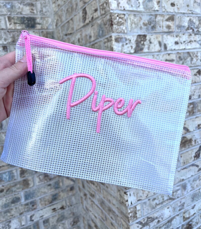 K&ouml;nnte beinhalten: Eine transparente, mit Rei&szlig;verschluss versehene Netztasche mit einem rosa Rei&szlig;verschluss und dem Namen "Piper" in Rosa gestickt. Die Tasche wird vor einer Backsteinmauer gehalten.