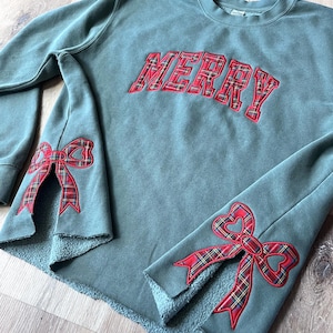 May include: A teal green sweatshirt with the word "MERRY" in red plaid lettering across the chest. The sleeves feature red plaid bow accents. The bottom hem is unfinished.
