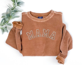 May include: A rust-colored, long-sleeved sweatshirt with the word "MAMA" in light gray, block letters across the chest. The sweatshirt has a ribbed texture and a crew neck. The sleeves are cuffed at the wrists. The background is white.