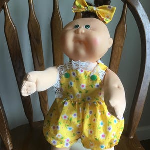 May include: A doll with a round face, green eyes, and dark hair tied in a topknot with a yellow floral bow. The doll wears a yellow romper featuring a white, blue, and pink floral pattern, white lace trim at the shoulders, and two green buttons.