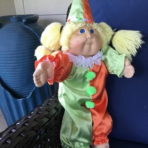 May include: A doll with blonde yarn hair in pigtails wearing a satin clown costume featuring a split orange and lime green color scheme, a matching pointed hat, a white ruffled collar, and three small green pom-poms down the front of the outfit.
