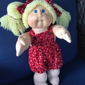 May include: A soft-sculpture doll with pale skin, bright blue eyes, and light yellow yarn hair styled in two pigtails. The doll wears a red romper featuring a pattern of black and white polka dots, accented with matching red polka-dot hair bows.