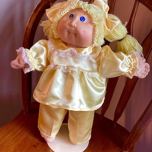 May include: A vintage doll with a round face, blue eyes, and yellow hair styled in pigtails. The doll is wearing a yellow satin pajama set with lace trim. It is posed on a wooden chair.
