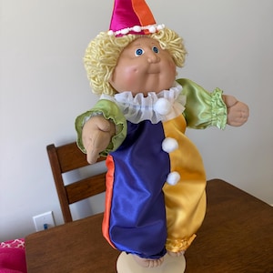 May include: A doll with blonde yarn hair wearing a satin clown costume featuring a purple, yellow, and orange color-blocked jumpsuit with green sleeves, a white ruffled collar, and a matching cone-shaped hat topped with a white pom-pom.