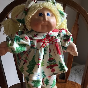 May include: A doll with blonde yarn hair styled in pigtails with matching bows, featuring blue eyes and dimpled cheeks. The doll wears a white long-sleeved top and pants set decorated with a colorful pattern of Christmas trees and a red ribbon bow.