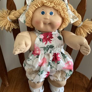 May include: A soft-sculpture doll with blonde yarn hair styled in two braided pigtails with matching floral fabric bows. The doll has blue eyes, a single visible tooth, and wears a white romper featuring a red and pink floral print.