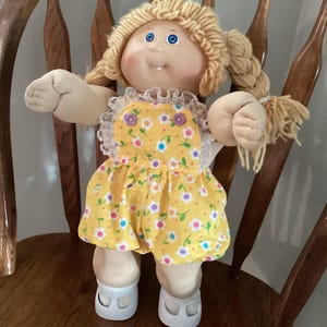May include: A vintage doll with blonde yarn hair styled in pigtails. The doll has blue eyes and a smiling expression. It wears a yellow dress with a floral pattern and white shoes. The doll is seated on a wooden chair.