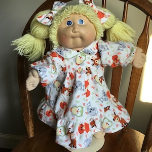 May include: A doll with light yellow yarn hair styled in two pigtails with matching bows. The doll has blue eyes, a round face, and wears a white dress featuring a repeating pattern of cartoon owls, foxes, and raccoons with small red leaves.