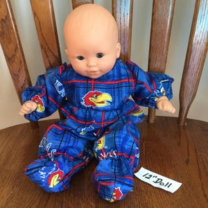 May include: A 12-inch doll wearing a blue plaid outfit with red accents and a yellow and red bird logo. The doll has a bald head and is sitting on a wooden chair.