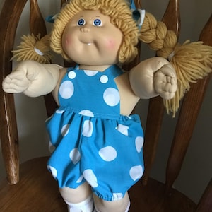 May include: A doll with light skin, blue eyes, and yarn hair styled in two blonde braids with blue and white checkered bows. The doll wears a bright blue romper featuring large white polka dots, white buttons, and white shoes with cutouts.