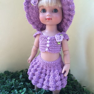 Può includere: Un completo per bambola in crochet viola con un cappello abbinato e decorazioni floreali. Il completo comprende un top corto e una gonna.