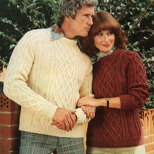 May include: A couple wearing matching cable knit sweaters. The man's sweater is cream coloured and the woman's sweater is burgundy. The man is wearing plaid trousers and the woman is wearing beige trousers. The image includes the text "SUNBEAM 285 Aran 86-107 cm bust 34-42 ins"