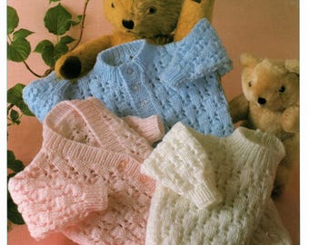 May include: Three knitted baby cardigans in pink, blue, and white. The cardigans are made with a delicate lace stitch and have button closures. The cardigans are displayed with two teddy bears.